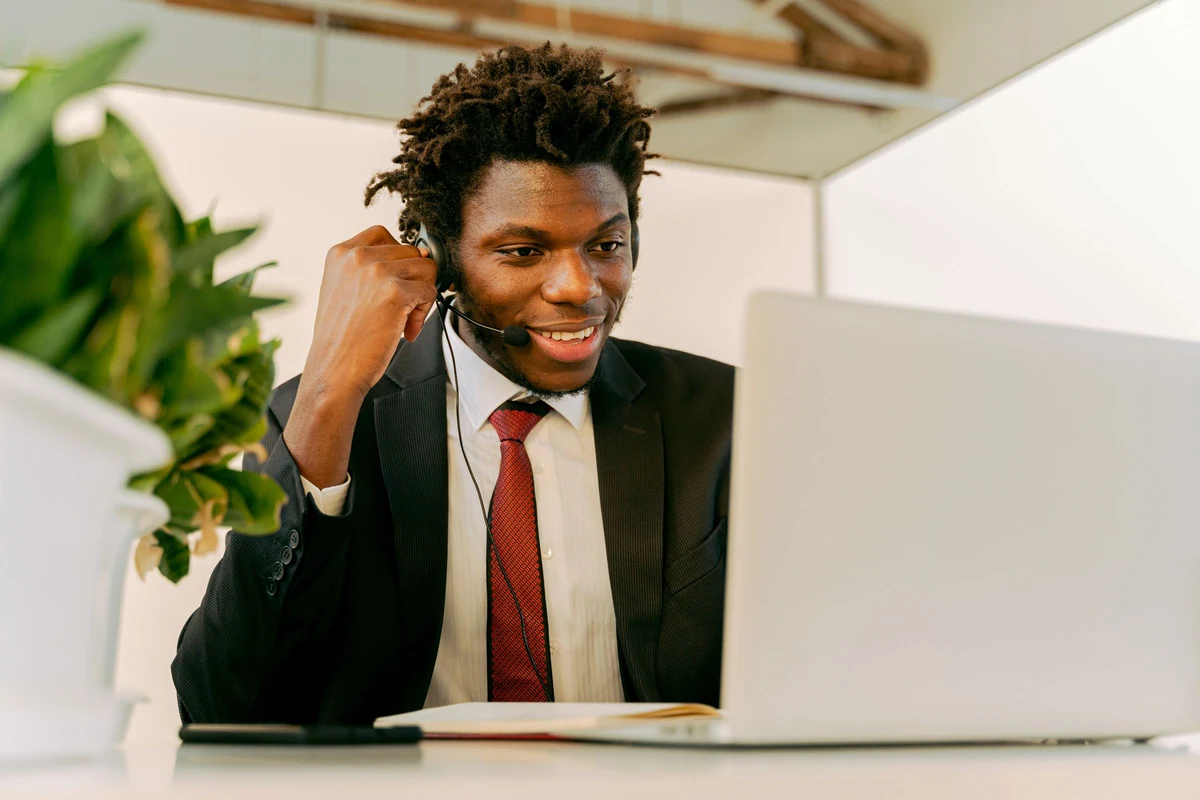 Comercial joven con auriculares y microfono sonriendo durante una videollamada frente a su portatil en la oficina