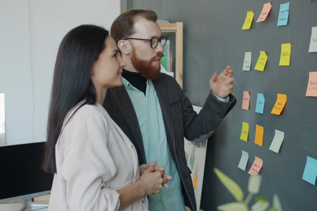 Dos profesionales comentando notas adhesivas de colores en una pizarra de una oficina moderna