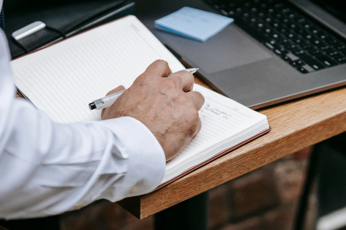 Manos de un profesional anotando en una libreta junto a un portatil abierto sobre el escritorio