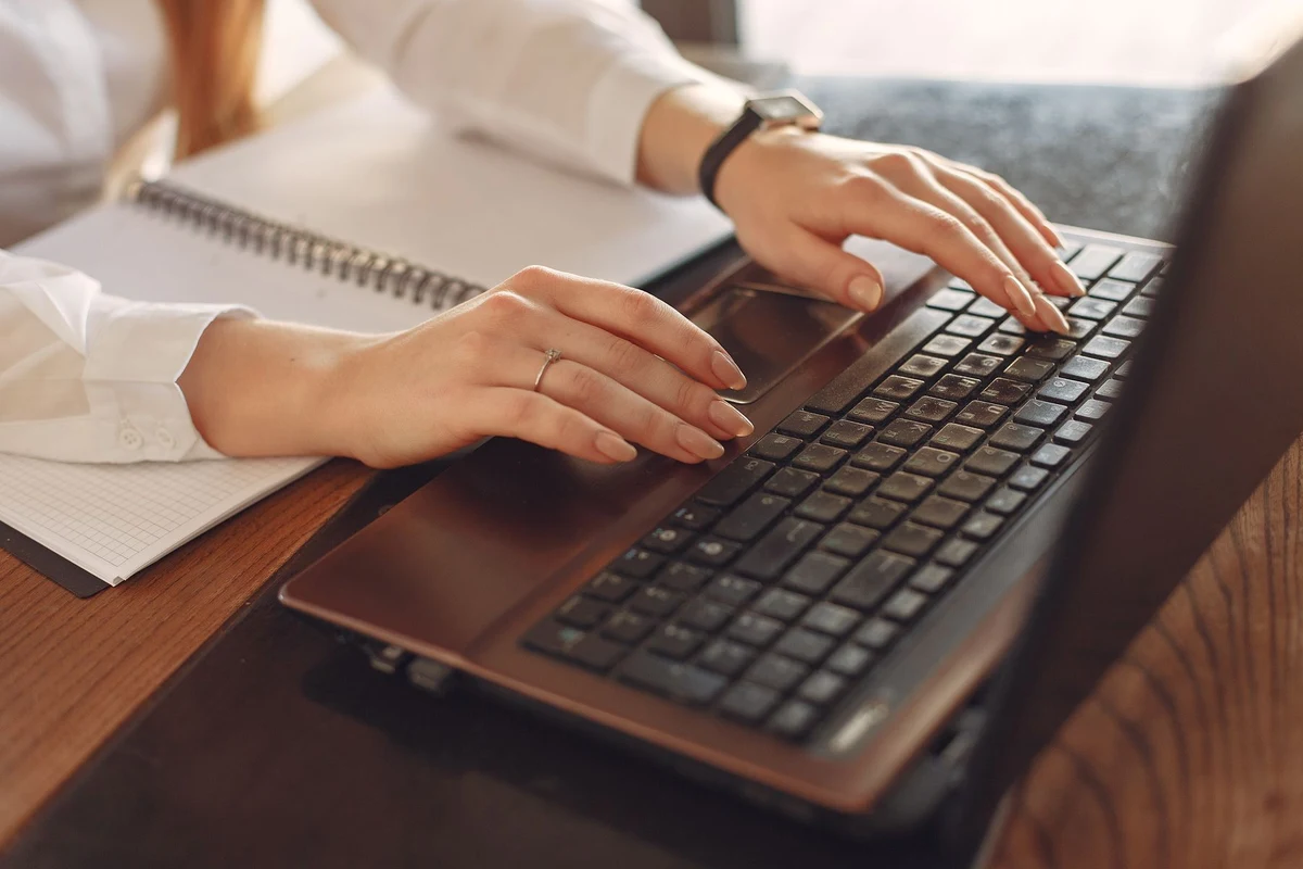Manos escribiendo en el teclado de un portátil junto a un cuaderno abierto