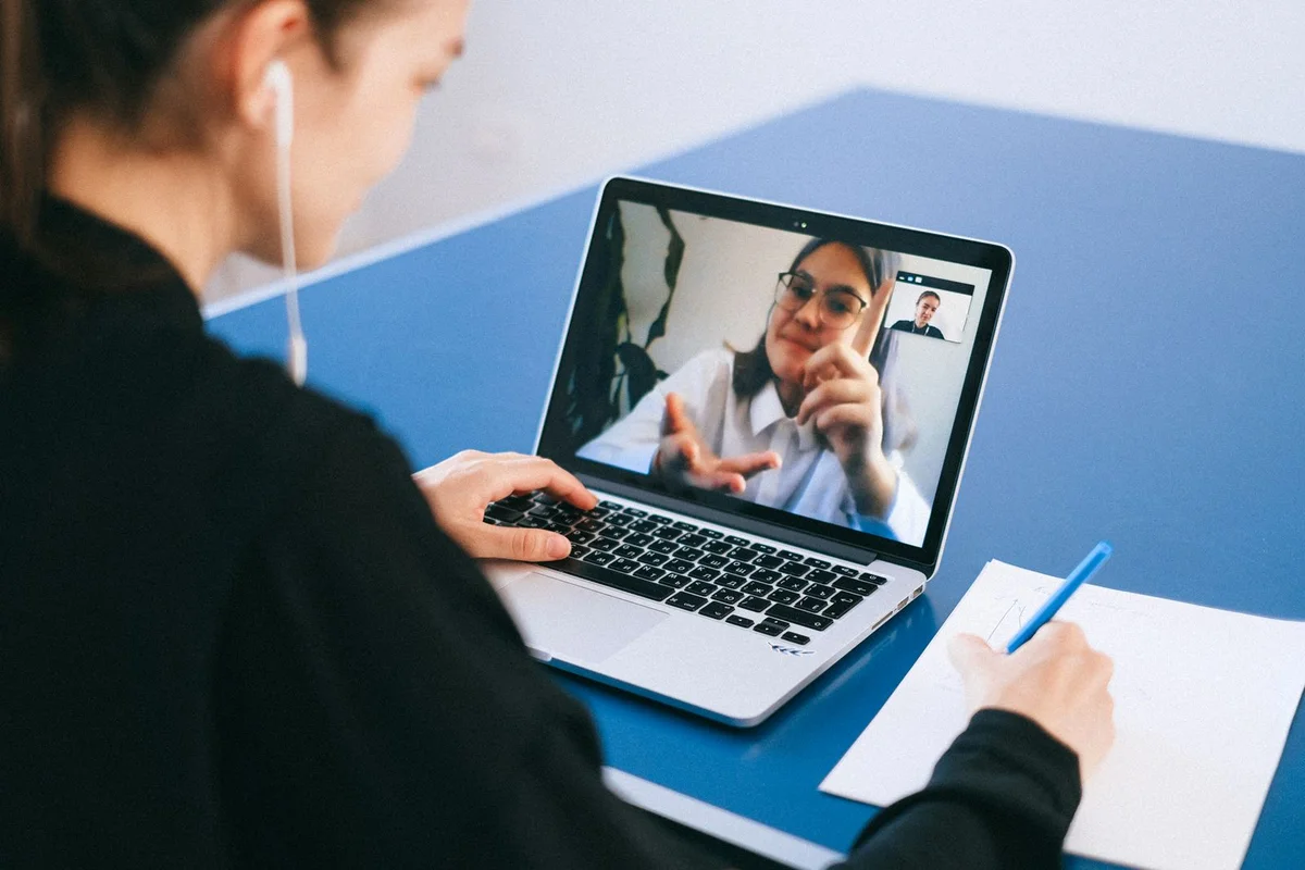 Persona con auriculares en una videollamada de trabajo frente al portatil mientras toma notas a mano